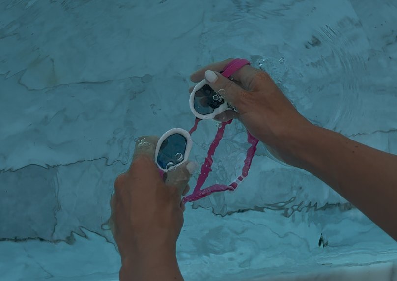 Both hands holding a pair of goggles just under the pool surface to wet the inside of the lenses